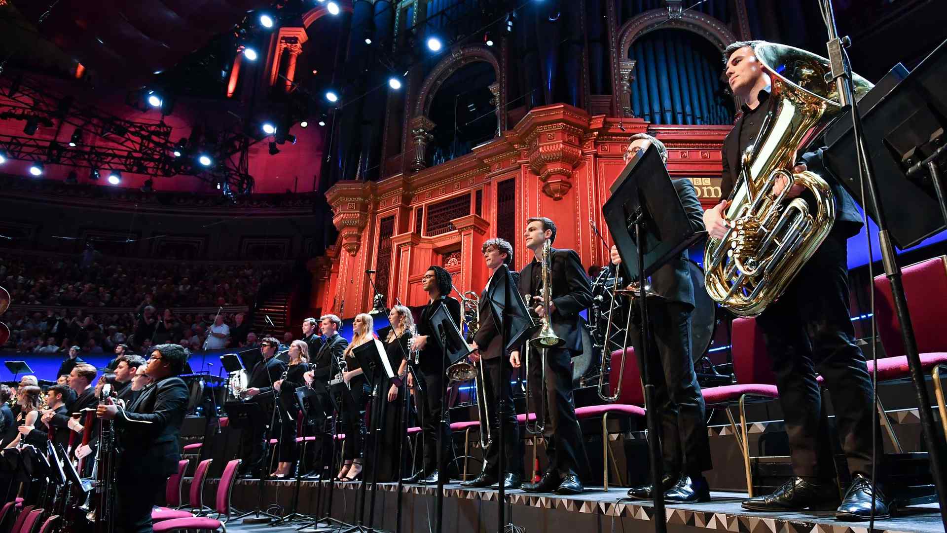 A row of Juilliard Orchestra musicians, including a tuba player, stand as they receive an ovation following a performance at the BBC Proms in the dramatically lit Royal Albert Hall