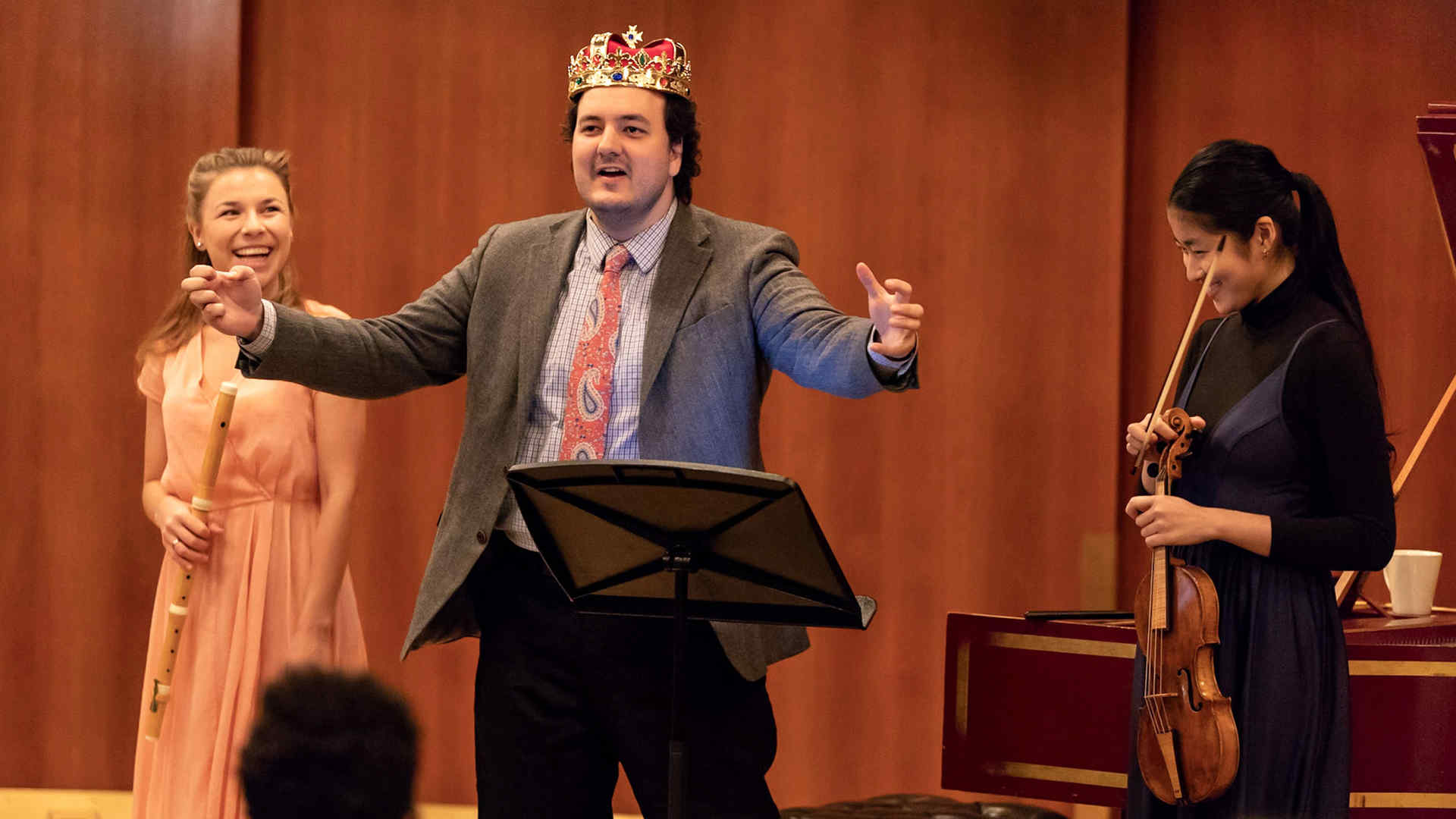 Carl, wearing a crown and flanked by Taya and Kako who are holding their instruments, speaking from the stage in Morse Hall