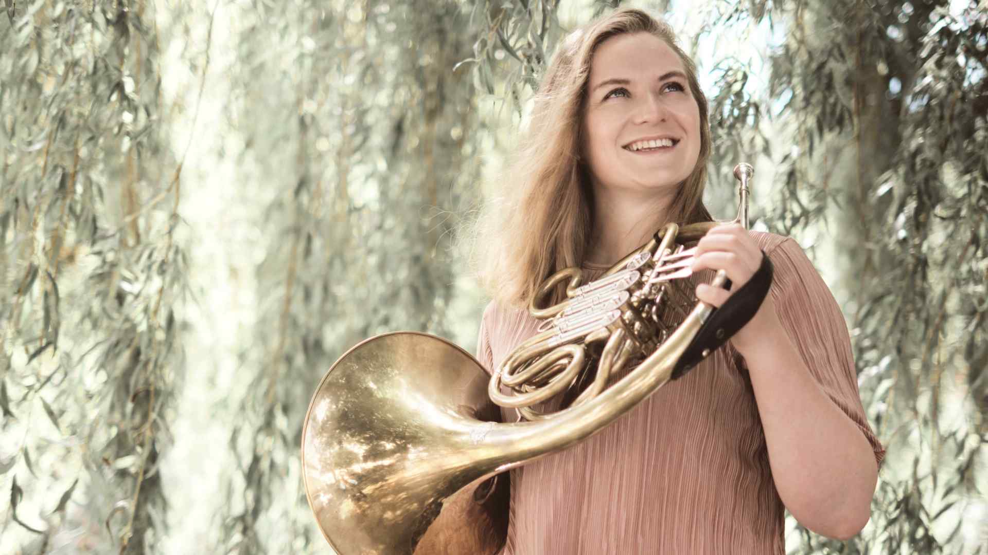 Headshot of Hannah Miller with her French horn shows her posing, smiling, in a sunny spot in front of a tree