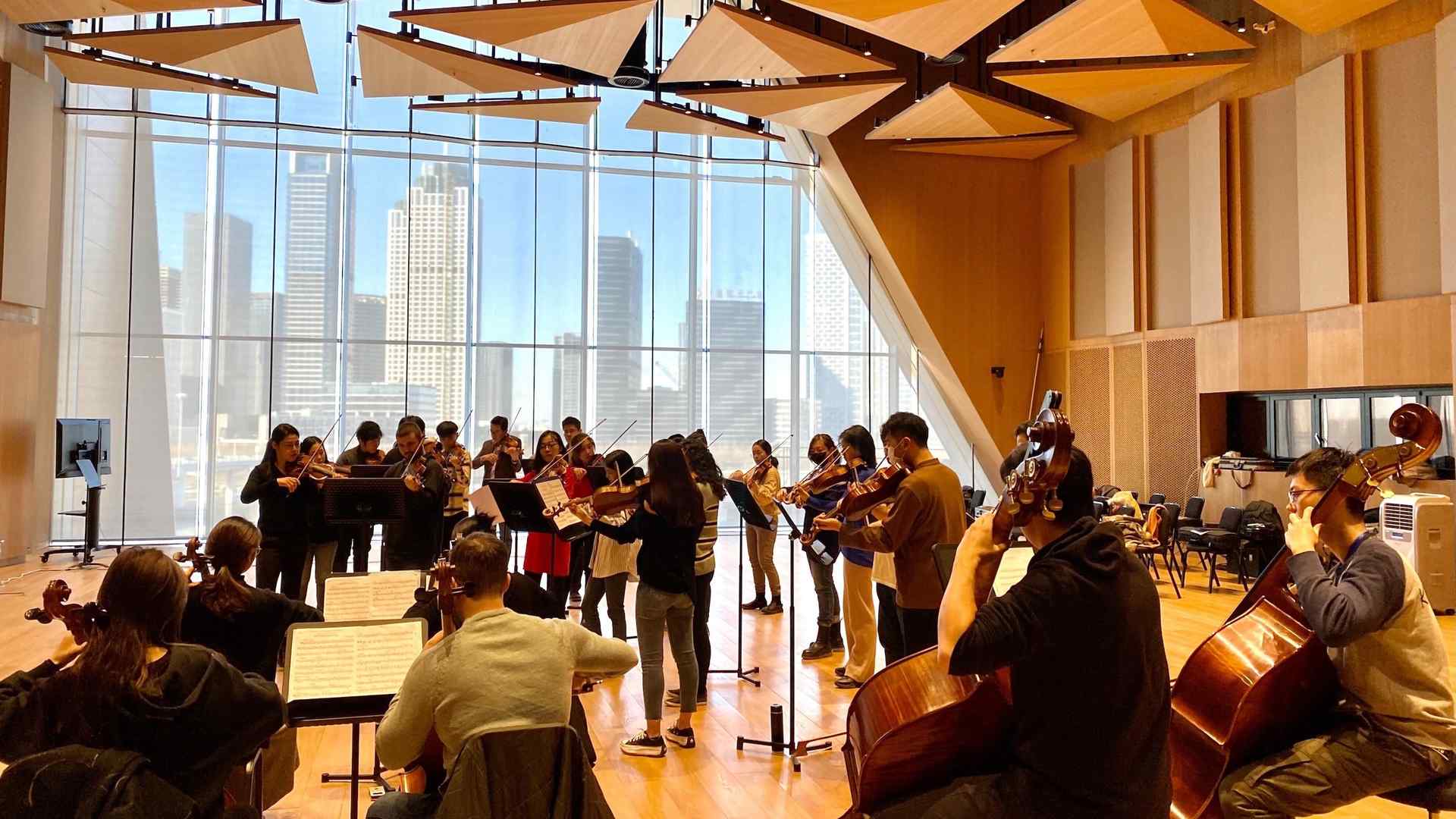 Orchestral musicians gathered in a rehearsal room featuring contemporary design and floor-to-ceiling windows looking out to skyscrapers against a blue sky