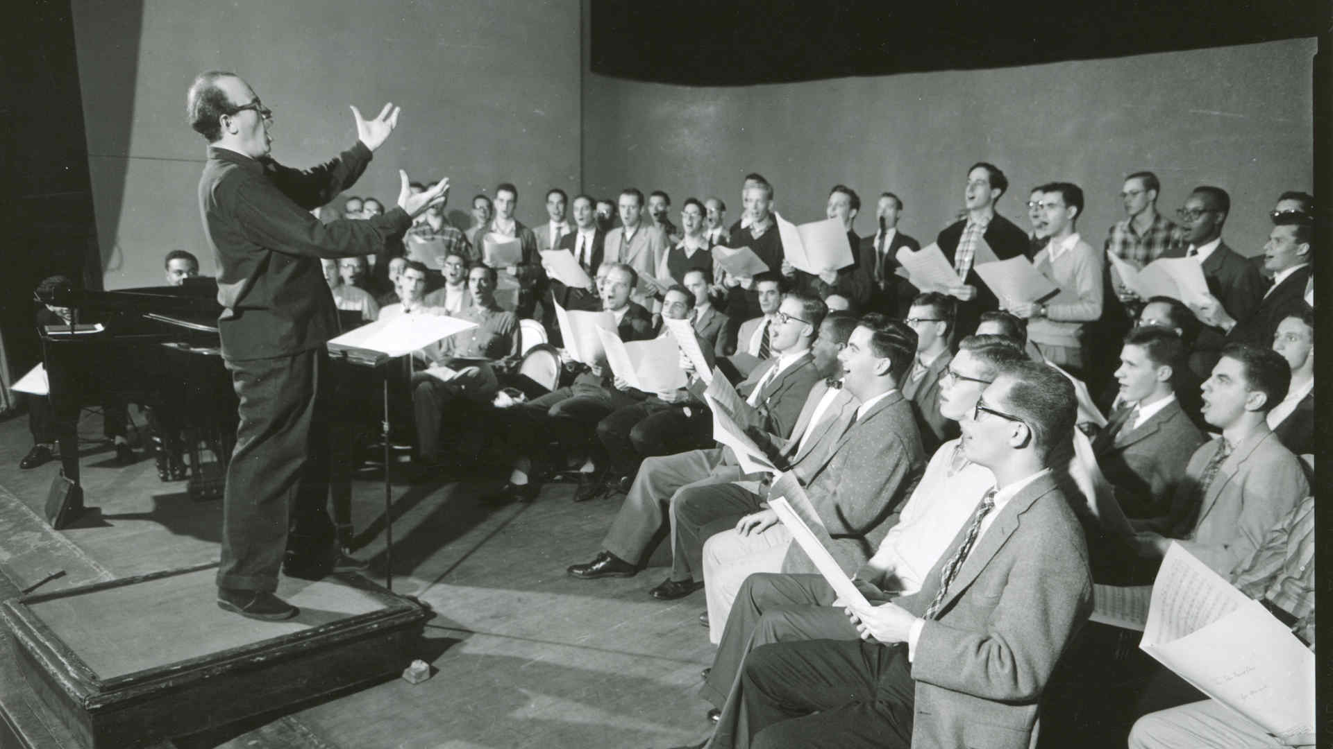 Archival (black and white) photo from a choir rehearsal of five rows of men in a semicircle. The conductor makes a passionate expression.