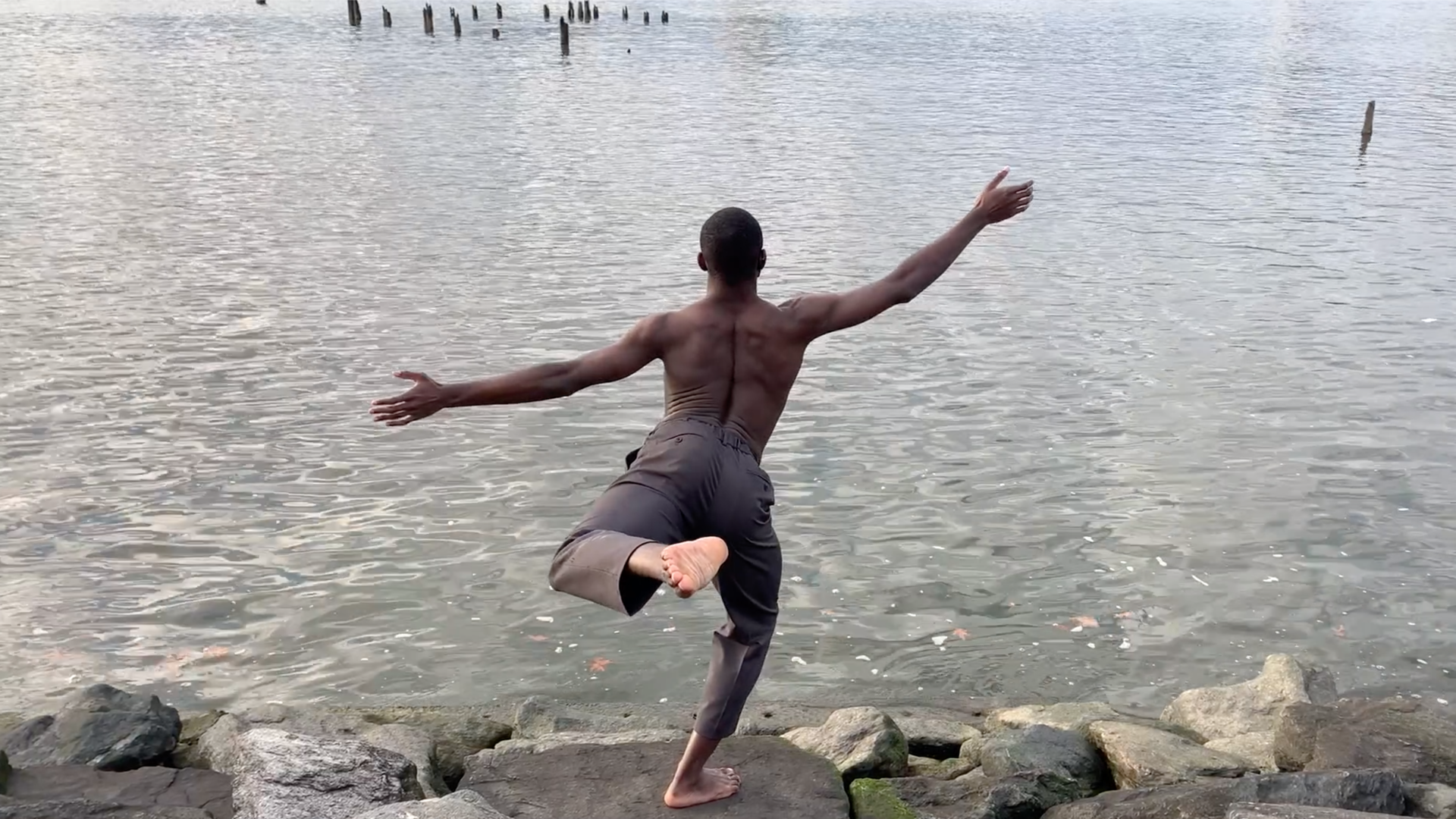 A dancer stands on the shore of a body of water, arms outstretched and with one leg raised, looking as though he might dive in