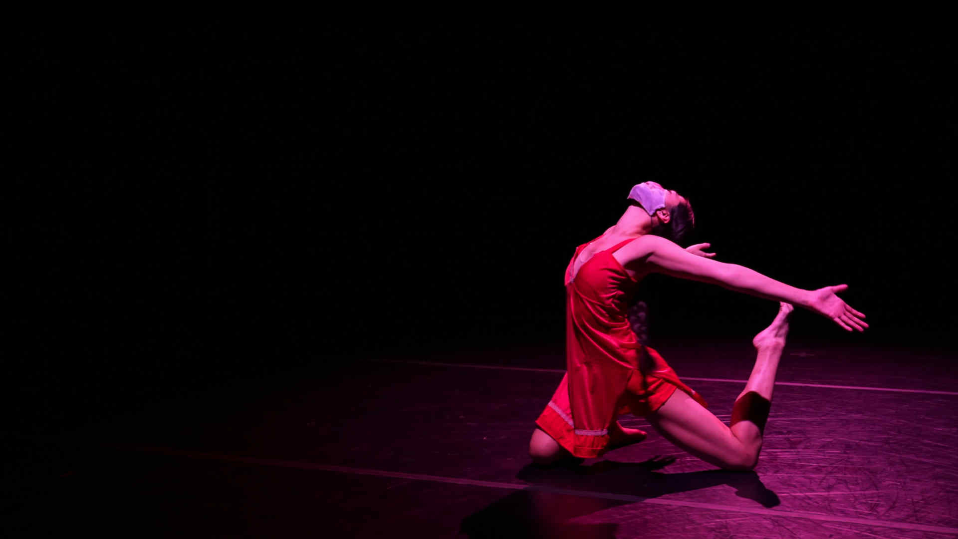 A dancer performing, costumed in a red dress in a kneeling pose with arms extended behind her