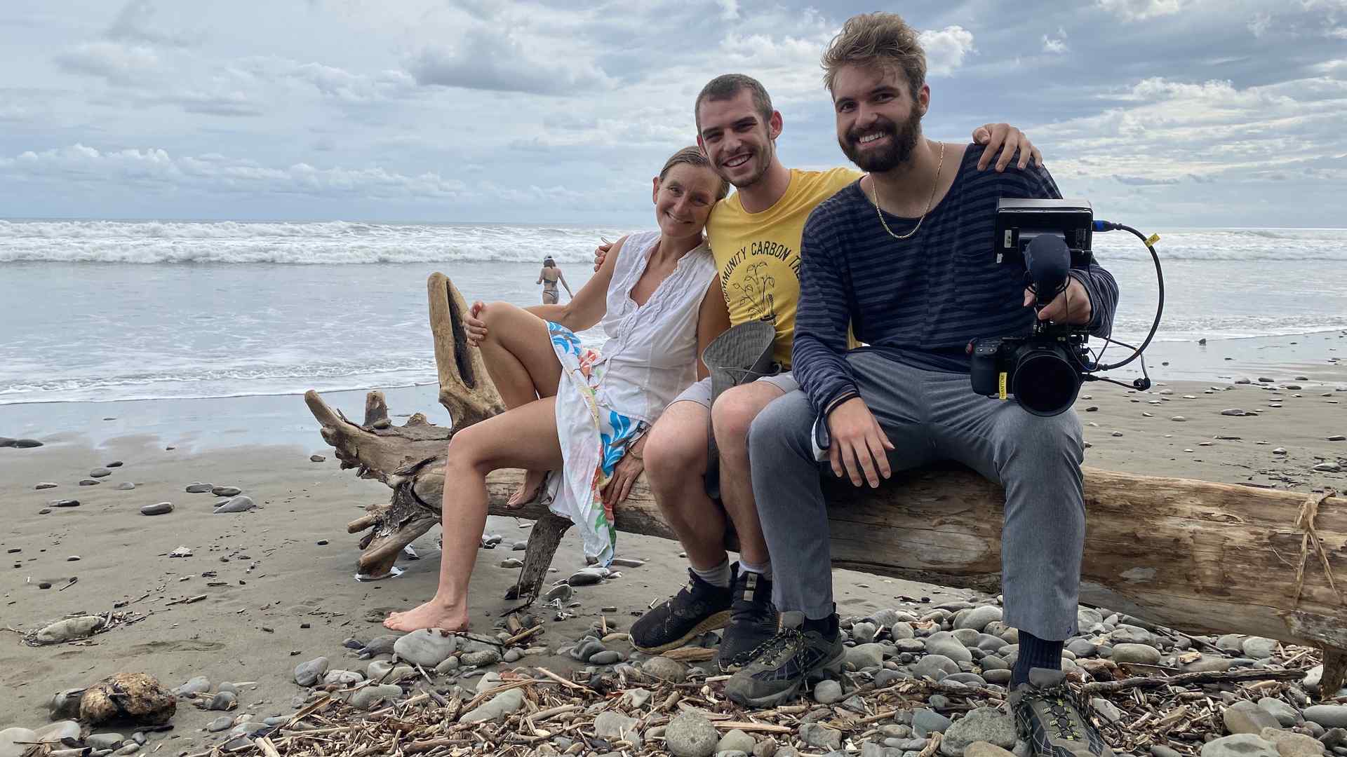 Jennifer Smith, Nathan Hirschaut, and Treyden Chiaravalloti at the shore. They are sitting on a large log of driftwood and Chiaravalloti holds a movie camera on his lap