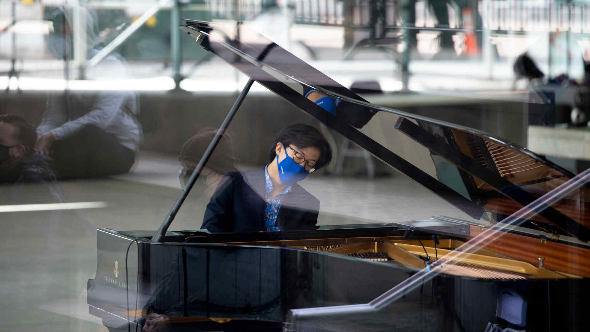 Pianist Derek Wang performing in the lobby of Alice Tully Hall, as seen through the exterior paned glass and passersby who gathered for the popup performance are visible in the reflection of the glass