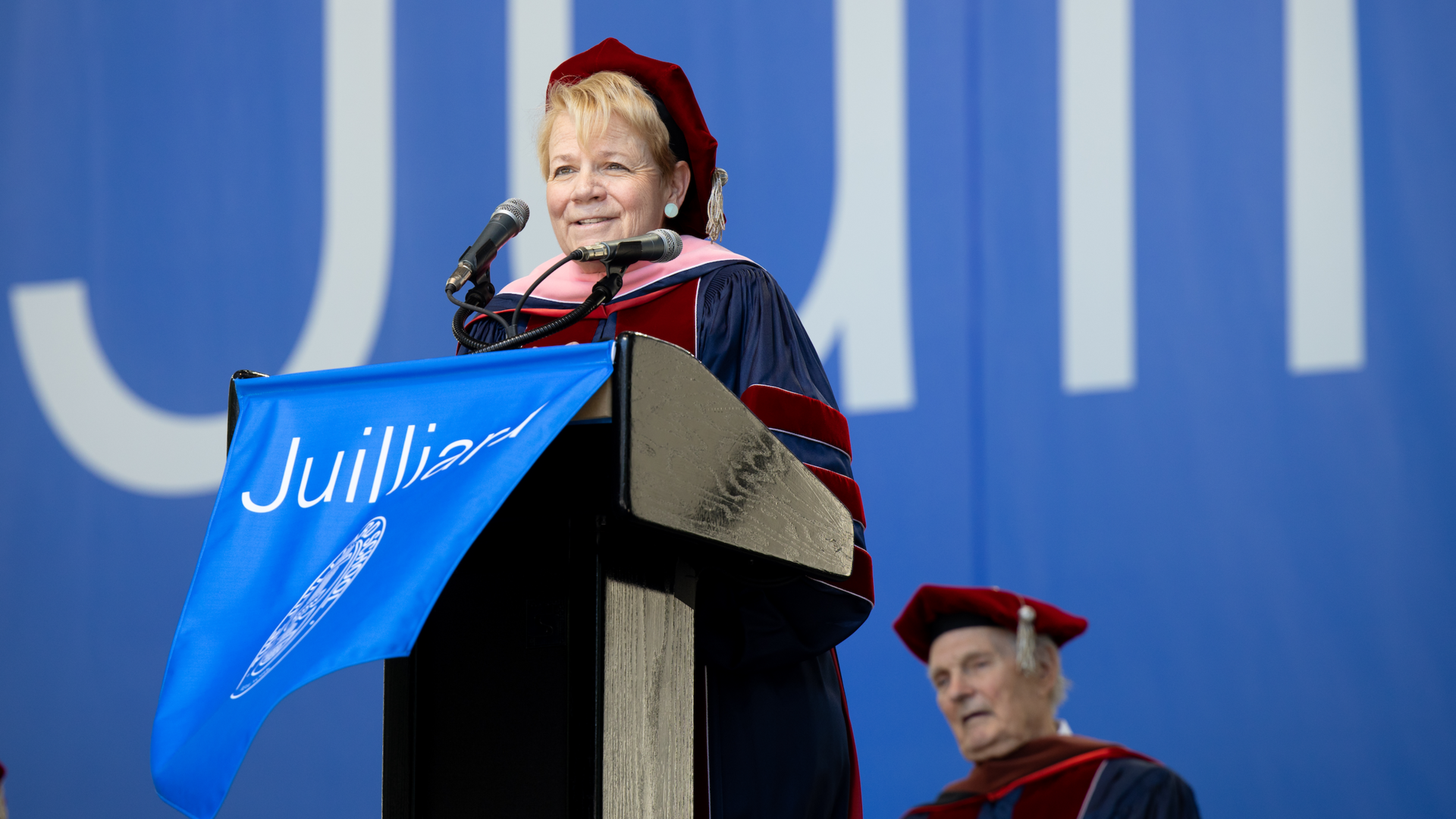 Marin Alsop in graduation regalia delivering a speech at a podium with a banner that says Juilliard and behind her is a big blue banner that says Juilliard