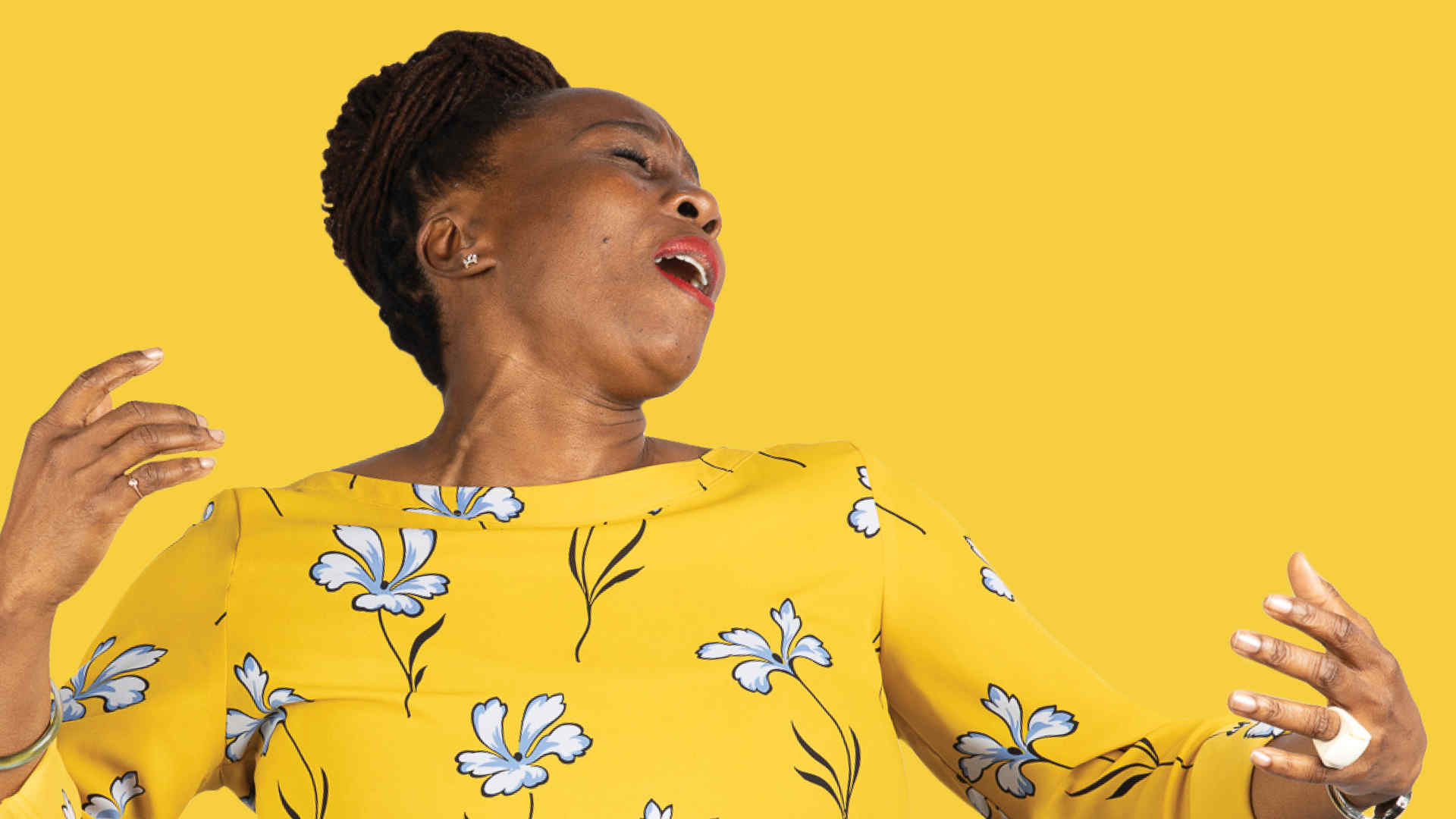 A woman wearing a yellow dress with flowers on it strikes an expressive pose and appears to be singing. The background is yellow.