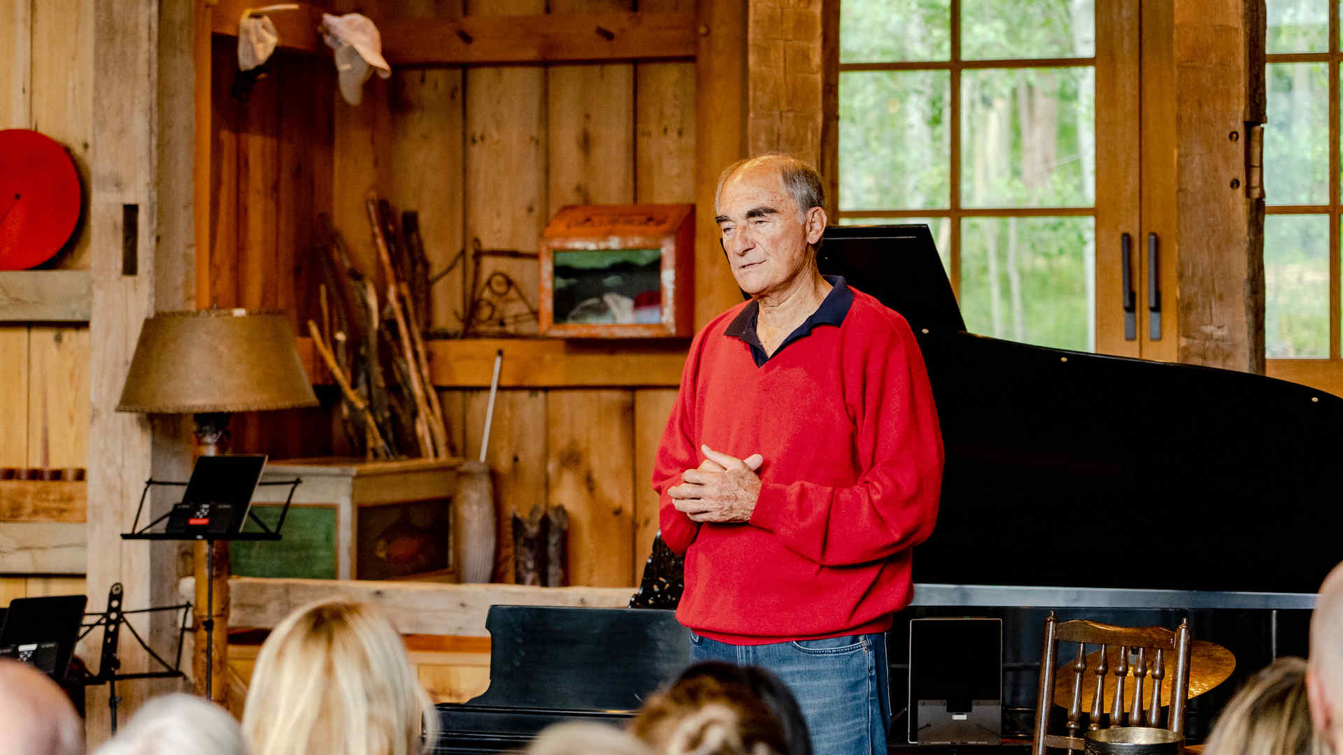 Vincent Mai, wearing a read sweater with blue polo shirt underneath and blue jeans, speaks to an audience. Behind him is a grand piano with lid open. The setting appears to be wood-paneled farmhouse or country house.
