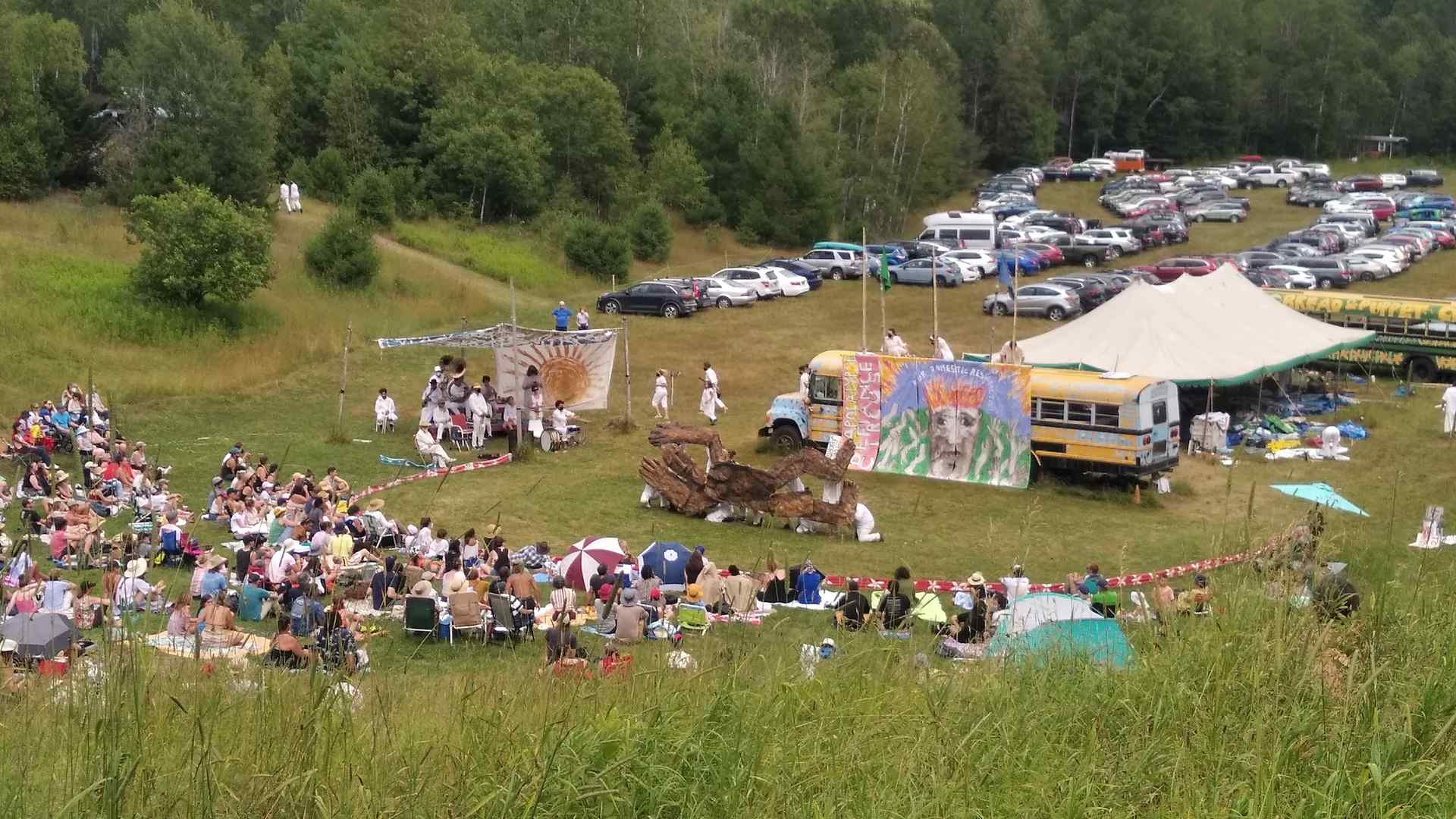 This image shows a grassy field, parked cars, and an audience seated in the grass, illustrating that Bread and Puppet takes place on a rural farm in Vermont