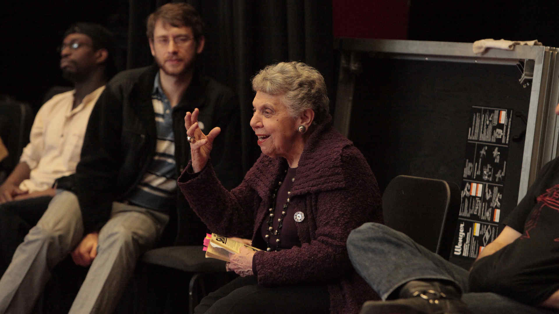 Eve Shapiro, seated, makes an emphatic hand gesture while teaching/directing and students, who sit on either side of her, look on