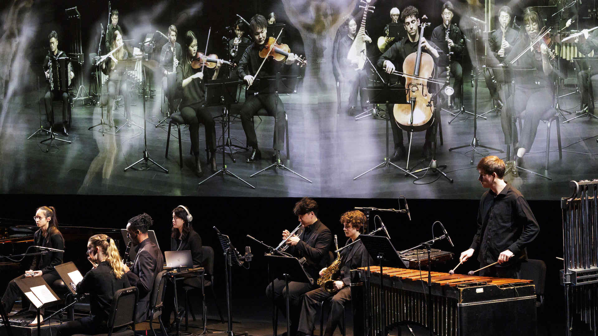 Photo from the interdisciplinary performance of 'In C' showing orchestral musicians and behind them on a screen are projections of musicians from the Tianjin Juilliard School as well as dancers