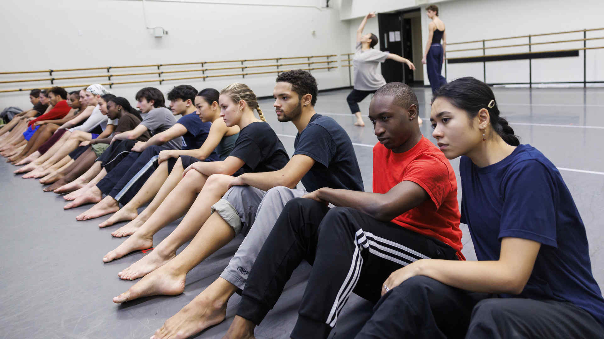 Dancers seated in a row on the studio floor during rehearsal