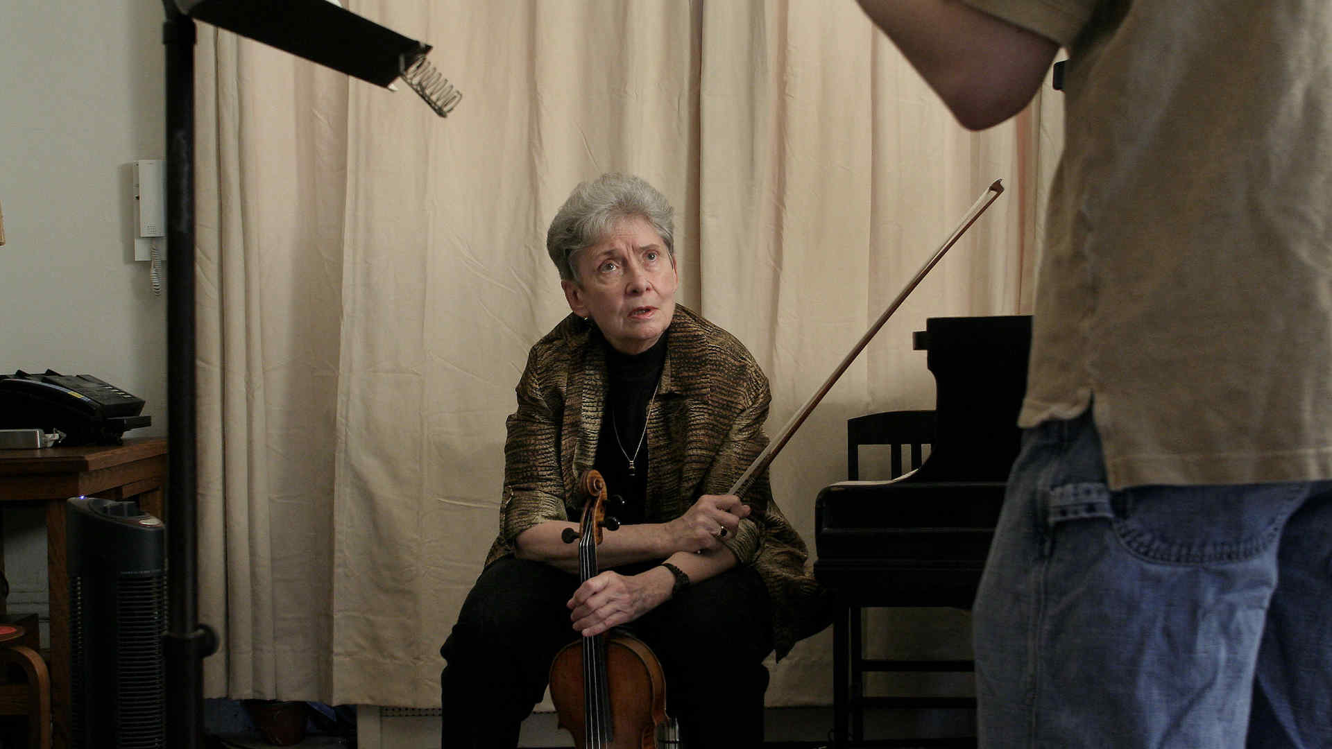 Sally Thomas is sitting with a violin and bow in hand, looking up at a student standing outside of frame. Thomas' facial expression is one of attention and perhaps inquiry or discussion. The room has a simple and functional decor, with a plain curtain backdrop and a music stand, which suggests a focus on music practice.