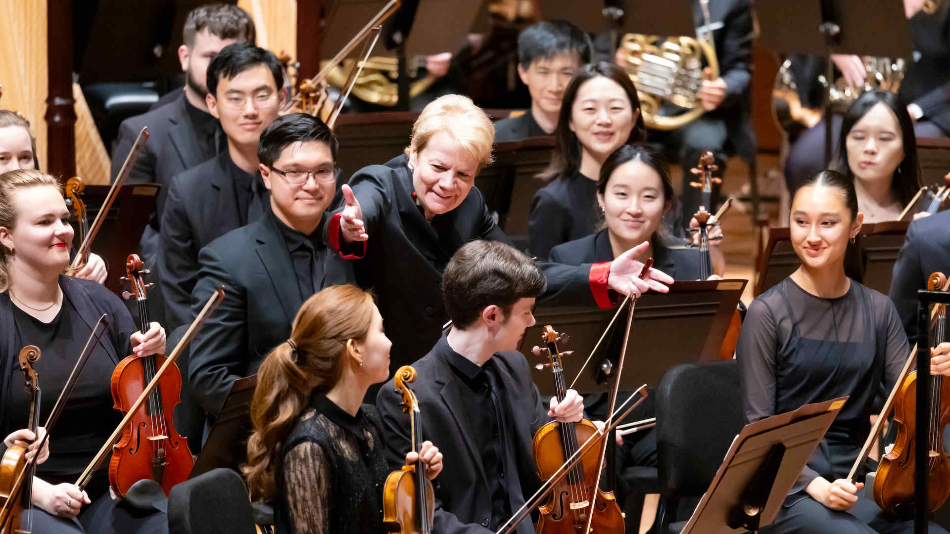 Alsop is engaging with the musicians of an orchestra, who are seated and holding their instruments, with smiles and attentive expressions on their faces.