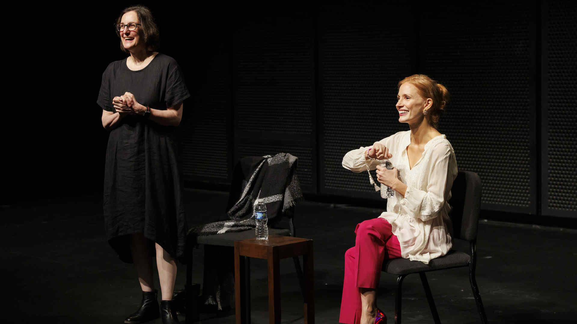 Jessica Chastain is sitting on a chair on stage, smiling and holding a water bottle. Next to her, Evan Yionoulis in a black dress is standing and clapping, also smiling warmly, during a Community Meeting at Juilliard.