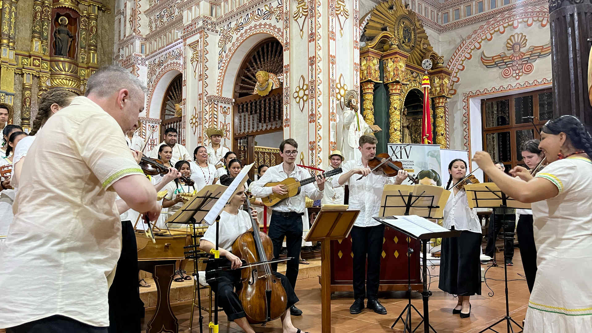 A group of musicians is performing in an ornate, historic church, with detailed carvings and religious figures visible in the background