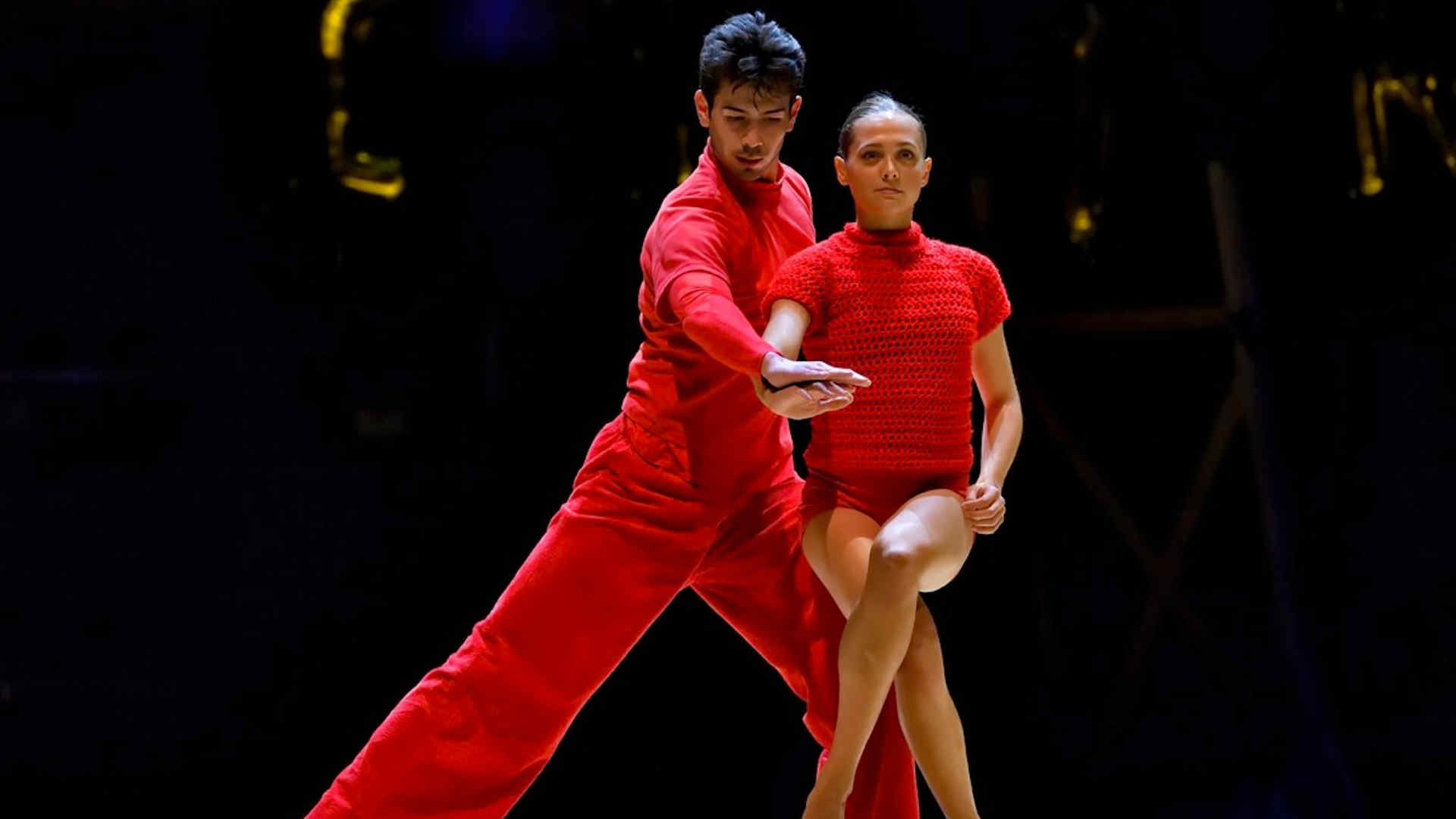 Two dancers perform on stage, both dressed in vibrant red outfits. One dancer is in a low lunge, extending their arm forward, while the other dancer stands beside them with one leg crossed over the other, conveying intensity and focus