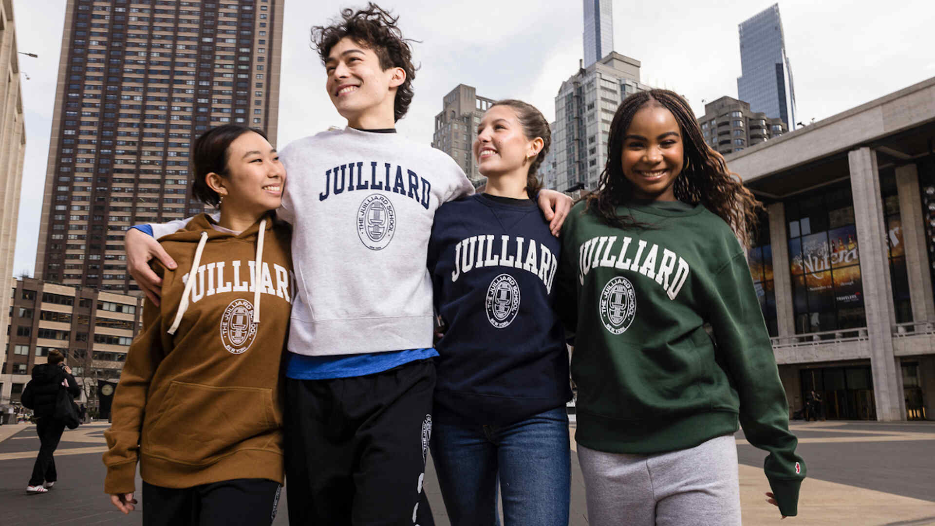 Four smiling students wearing Juilliard sweatshirts in different colors, standing arm-in-arm outdoors