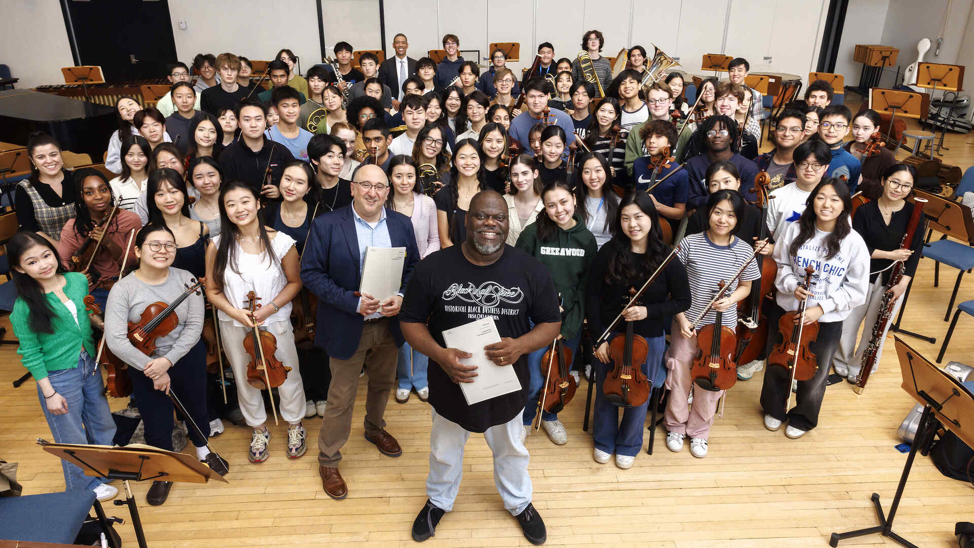 A group photo with Jeff Scott standing front and center with members of the Juilliard Pre-College Orchestra surrounding him. The group of young musicians is smiling and gathered in a rehearsal room with music stands and chairs in the background