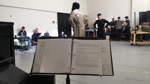 A music stand holding a script in foreground, actors rehearsing in background