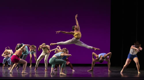A dancer leaps through the air while other dance and there is a purple background