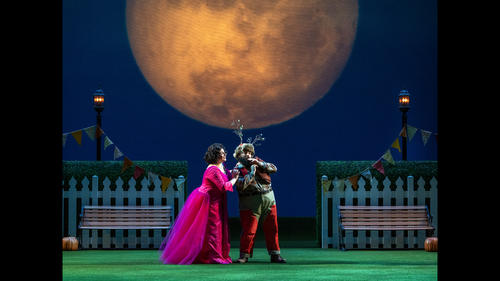 Two singers in a garden beneath a large orange moon in a production photo