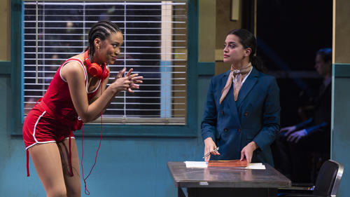 Two actors mid-performance in a scene, standing in an office. One appears to be trying to convince or ask the other for something.