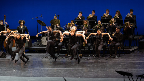A vibrant performance featuring a group of dancers in black, sequined costumes, energetically executing a dance routine on stage. Behind them, a big band with brass instruments provides live music, creating a dynamic backdrop for the dancers. The setting suggests a collaborative concert or musical performance, highlighting the intersection of live music and dance.