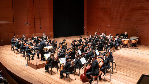 The image depicts The Juilliard Orchestra in performance, with musicians dressed in black attire, seated on the wooden stage at Alice Tully Hall and playing their instruments. Conductor Jörg Widmann is leading the ensemble. The stage is well-lit, highlighting the orchestra's focus and coordination during the performance.