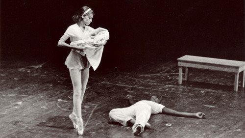 The archival (black and white) image depicts a ballet performance with a dancer in mid-pose, standing on pointe with one leg lifted slightly, holding a bundle which suggests a baby. She is looking down at a dancer lying on the floor beside her, creating a dramatic scene. In the background, there's a simple wooden bench on an otherwise empty stage, highlighting the stark emotional moment between the dancers