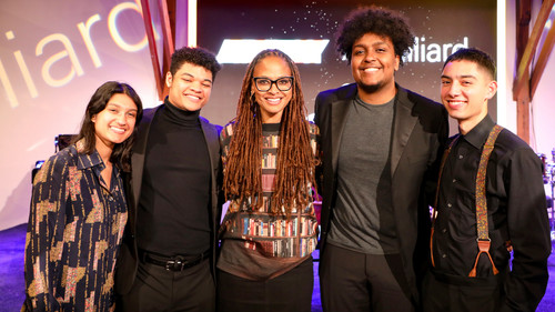 This image features a group of five individuals posing together for a photo. They all appear to be cheerful and are standing in front of a backdrop that includes the word "Juilliard" in large, stylized letters, suggesting that they are at a special event.