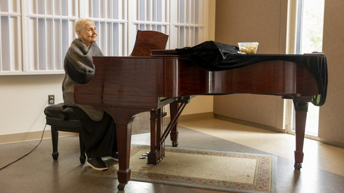 Marlena Malas seated at a grand piano in a room with a large window providing natural light. Malas is wearing a gray shawl over her shoulders and appears to be either in a moment of rest or contemplation. The piano's lid is open. There's a sense of calm and poise about her, indicative of a seasoned musician and teacher.