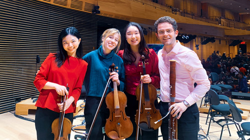 Four smiling musicians posing together on a concert hall stage. They are holding string instruments: two violins, a viola, and a bassoon. The background reveals an empty stage with chairs and music stands, indicating a rehearsal or a performance break.