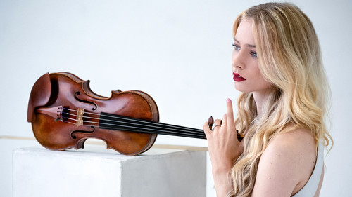 Mariella Haubs is pictured in profile, gazing thoughtfully into the distance. She rests her hand on the neck of a violin that sits on a white pedestal, suggesting a connection between her elegance and the grace of the instrument. The simple background focuses attention on the synergy between the musician and her violin, evoking a sense of both contemplation and readiness to perform.