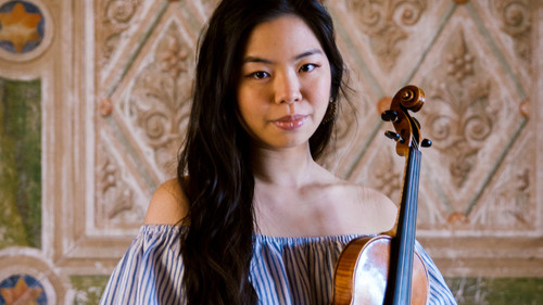Amelia Sie holds her violin , exuding a sense of calm confidence. She wears a casual, off-the-shoulder striped top and white pants, which gives her a relaxed but stylish appearance. Behind her is an ornate wall with intricate patterns, which adds an elegant contrast to her modern attire. Her expression is serene and contemplative, suggesting a deep connection with her instrument.