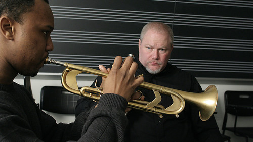 On the left, a student plays trumpet. On the right, Jaudes observes the student intently. The chalkboard in the background has musical staff lines on it, suggesting a music room or classroom designed for music lessons or practice.