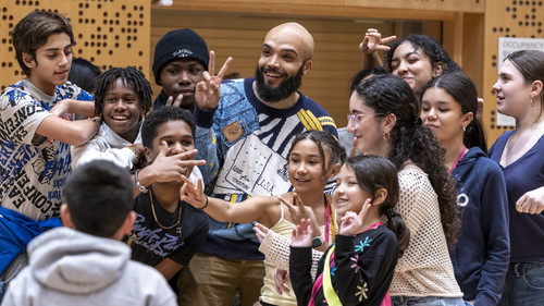 A joyful moment among young students gathered around Marcus Norris in an orchestral rehearsal studio at Juilliard. Norris is smiling broadly and wearing a stylish, patterned sweater. He stands at the center, surrounded by the enthusiastic students posing playfully—some making peace signs, while others smile or gesture animatedly towards the camera.