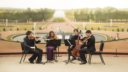 A string quartet performing in front of a large mural depicting a classical garden scene. The players are deeply engaged in their music. The setting, highlighted by the grand mural of a baroque garden with neatly trimmed hedges and statues, adds a formal and historic ambiance to their performance. The musicians, dressed in concert attire, enhance the elegant and artistic atmosphere of the scene.