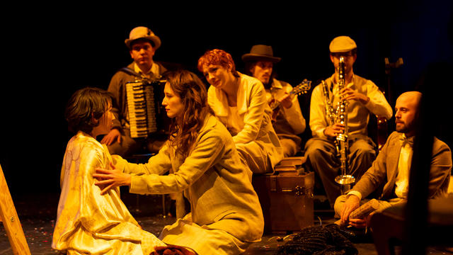 A group of actors sits in a circle playing musical instruments