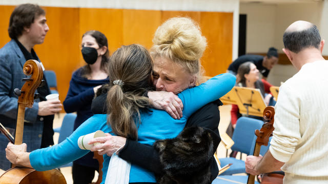 Two individuals embrace emotionally, reflecting the concept of reunion, in a crowded orchestral studio. The photo appears to have been taken before or after a rehearsal.