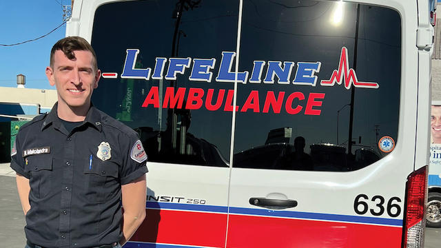 Nathan Makolandra dressed in a uniform standing beside an ambulance. On the back window of the ambulance is visible the logo "LifeLine Ambulance," which is the ambulance company Makolandra works for.