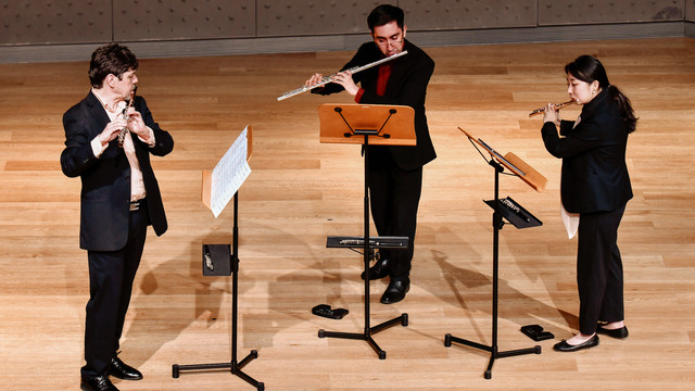 Three instrumentalists, dressed in performance black, are standing on stage in a semi-circle performing