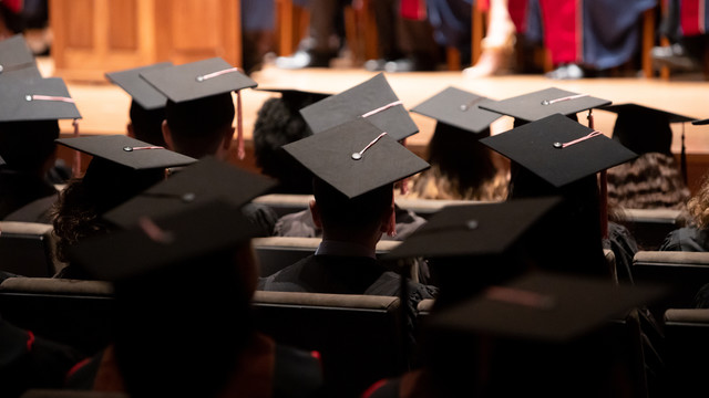 A group of graduates during a commencement ceremony. They are wearing traditional black caps and gowns. The setting is Alice Tully Hall with rows of seating filled with the graduates. The angle of the photo suggests it’s taken from the back of the hall, focusing on the back of the graduates' heads and their mortarboards, with one in the foreground serving as a focal point.